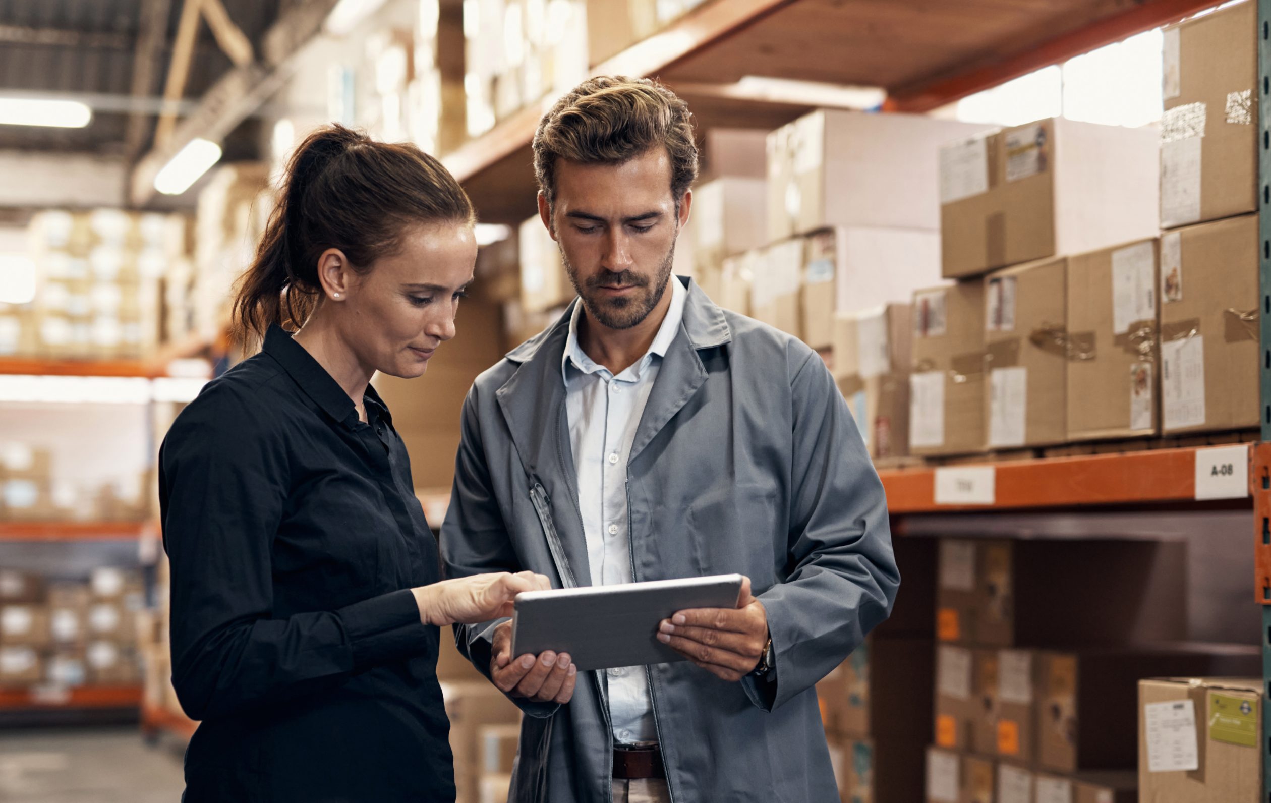 Photograph of two people looking at a tablet while standing in front of warehouse shelves filled with boxes