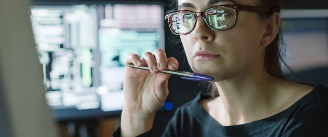 Photo of woman with glasses looking at a computer monitor while holding a pen.